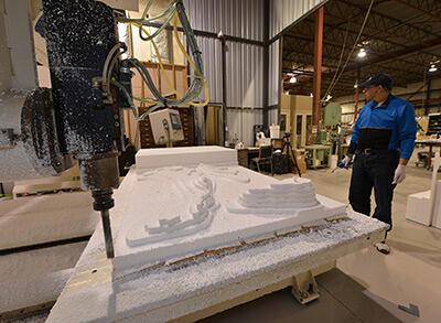 A worker standing beside a special milling/cutting machine, at work carving a copy of a bone onto a huge slab of foam.