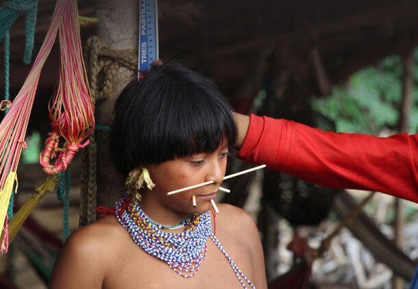 A young person with a dark bowl cut, a stick through their nose and facial decoration on the chin, and an elaborate beaded necklace.