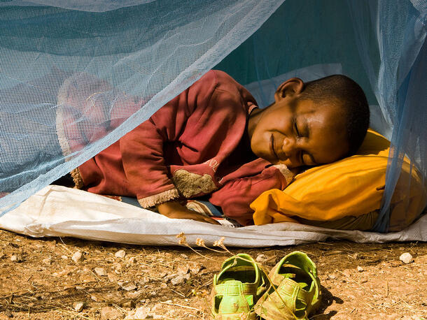 A child sleeps on a mat on the ground surrounded by an insecticide treated bed net.