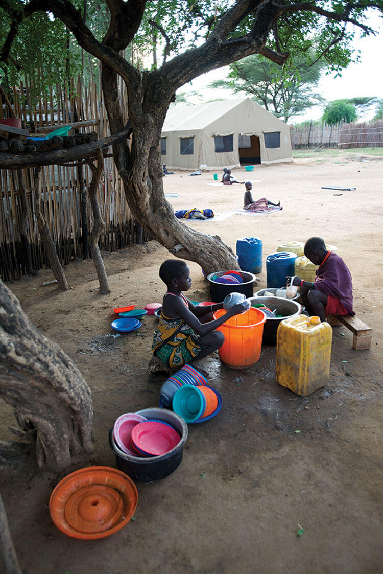 Two young people sit or crouch on the ground and wash colorful dishes with water and soap in buckets.