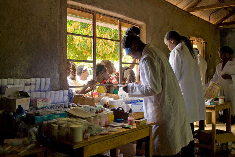 Four adults in lab coats stand inside a room where a table is covered in medicines as a crowd of people stand outside at the window.