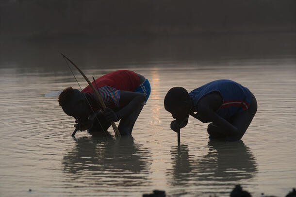 Two people stand in water and crouch to drink from plastic drinking tubes.