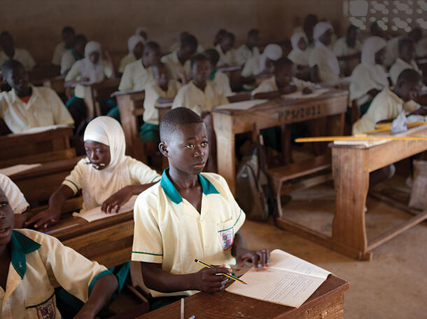 Abdulai Iddrisu sits in the front row of a classroom with over 20 other students and holds a pencil over an open notebook.