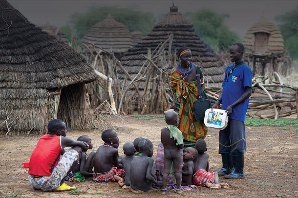 Paulo Lovul and Aleper Akol Naparinga stand in front of a seated group of children and an adult and hold out a flip chart to education about guinea worm.