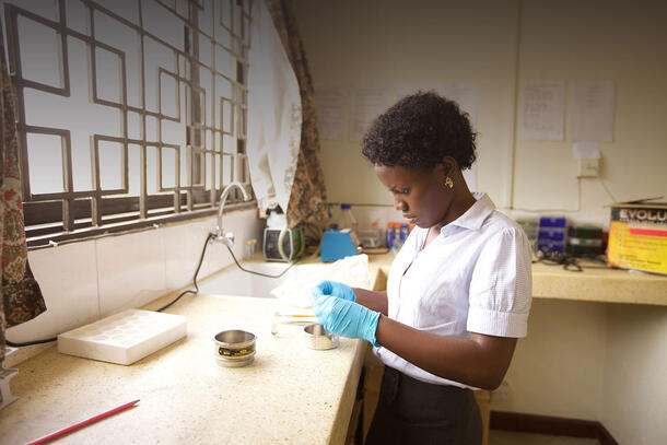 A laboratory technician stands at a counter beside a sink wearing plastic gloves and works to put something in a small circular bowl.