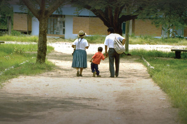 A child, Luis Fermin Tenorio, walks between his parents on a dirt path between greenery and leans to the right due to effects of polio.