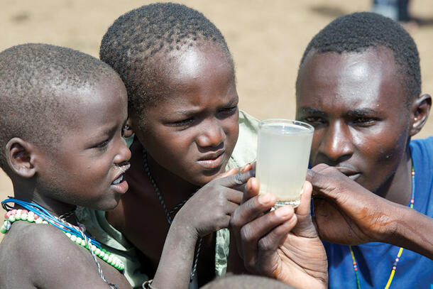 Emmanuel Ekadeli crouches beside two children and points out copepods in a glass of water to two children.