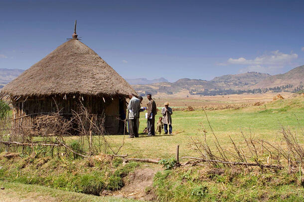 Two healthcare workers, one with a clipboard, speak to an adult who stands beside a child, an older person, and a hut with a woven roof.