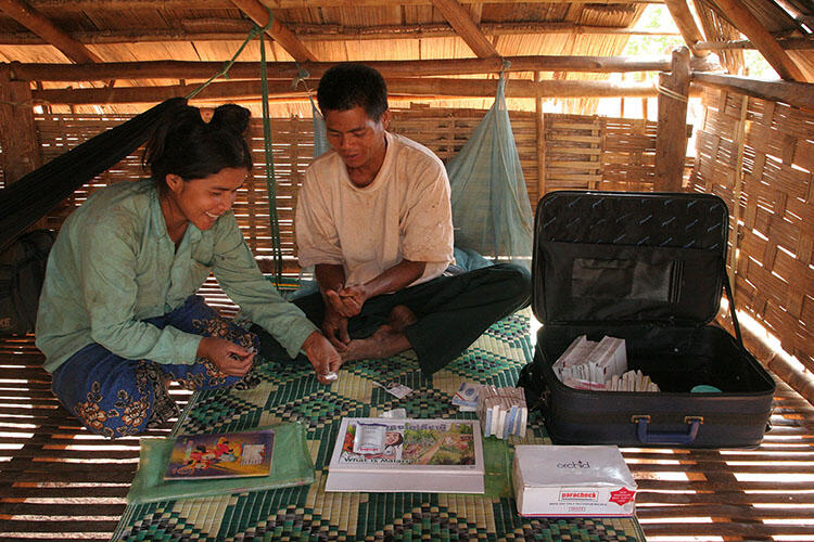 Health worker Tek Phon sits on the floor of a woven hut with another person and shows them a Rapid Diagnostic Test for malaria.