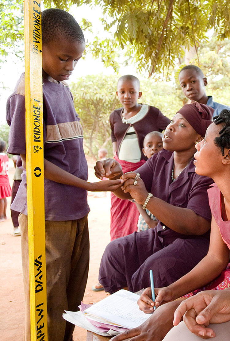 A seated adult holds a standing child's hand and speaks to them while another adult takes notes.
