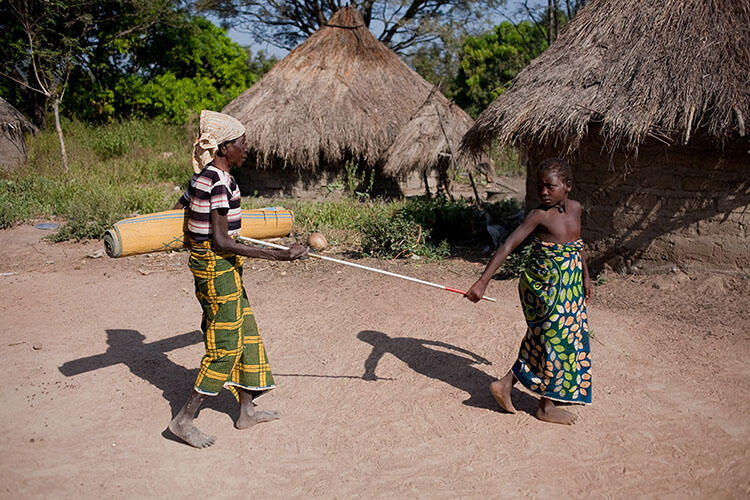 A child leads a blind adult using a long stick which they both grip as they walk past two huts.