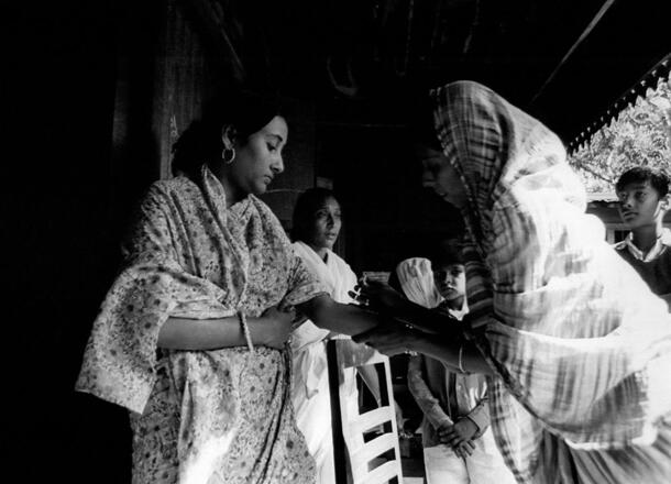 An adult in floral patterned clothing receives a smallpox vaccination via needle from a person wearing a light head scarf.