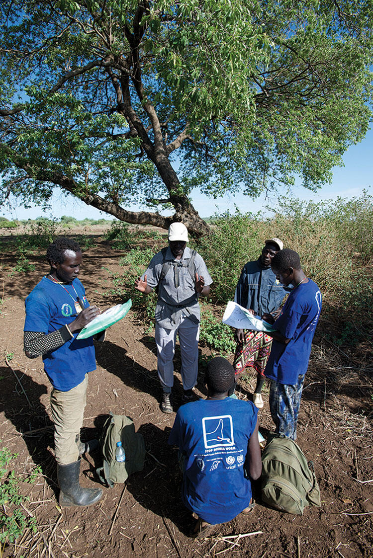 Makoy Samuel Yibi stands and speaks to a group of four adults, three of whom are wearing t-shirts from the Guinea Worm Eradication Program.