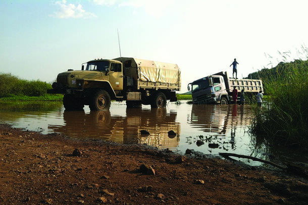 Two trucks traversing a muddy area in South Sudan. One of them is stuck in the mud slightly sideways, with four people standing around it.