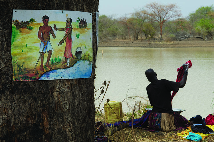 A person washes clothing on the banks of a dam. A colorful illlustration of a guinea worm patient is hung on a tree in the foreground.