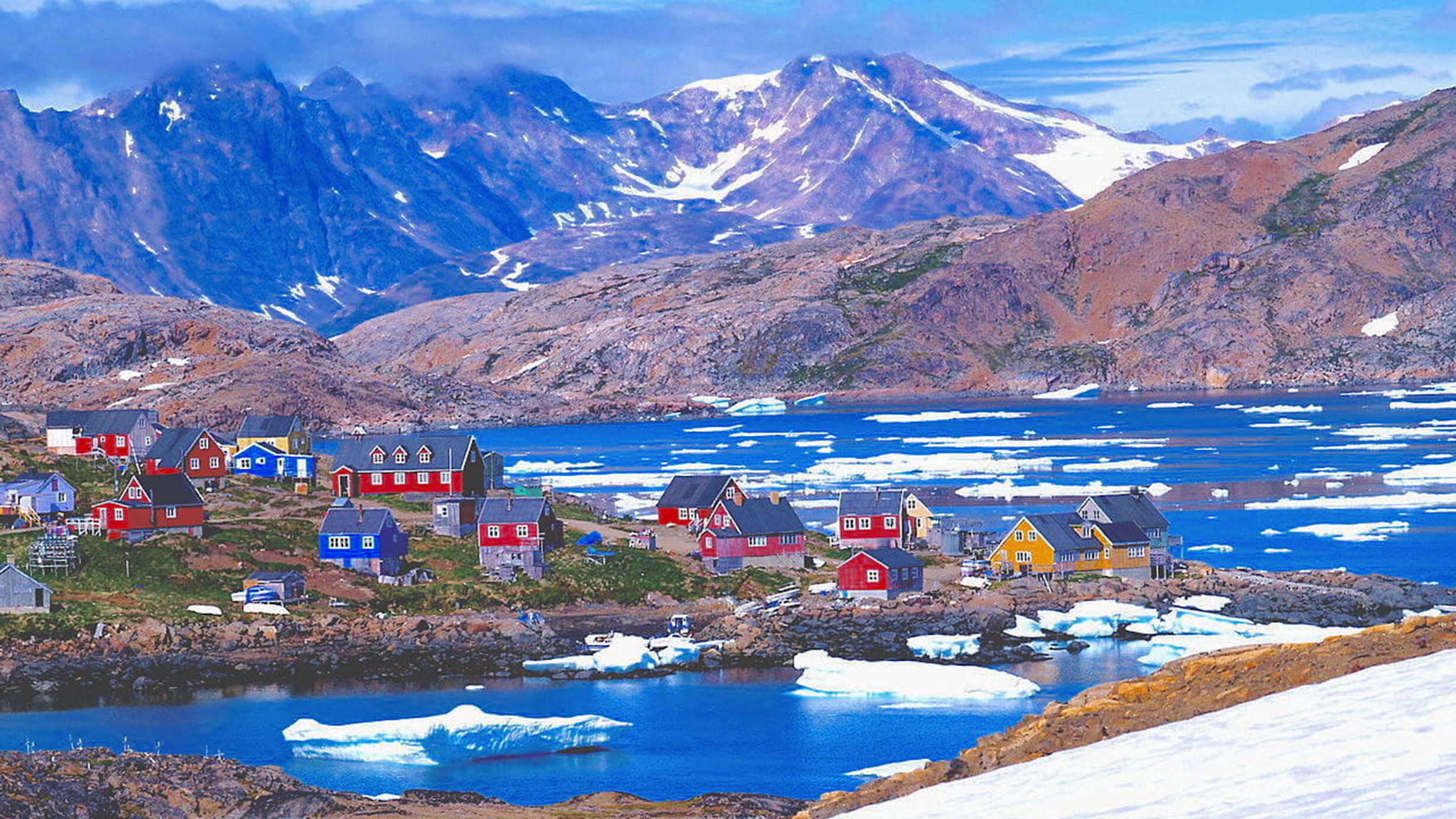 Colorful buildings on land surrounded by water and melting floating ice, plus mountains in the background, in the town of Tasiilaq in Greenland.
