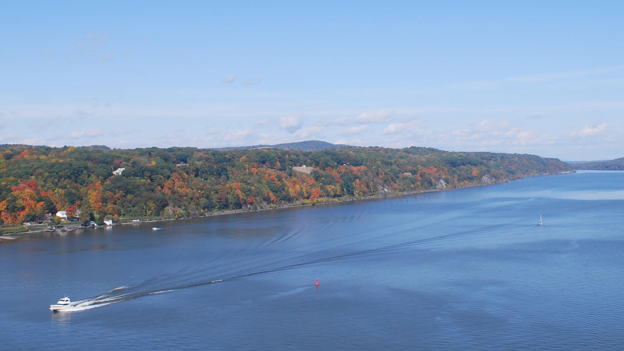A small boat making its way down the Hudson River with dense trees along the banks that have changed color for fall.