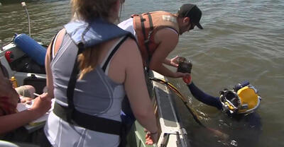 A scuba diver with their head out of water hands an object to someone in a motorboat. Two more people stand and sit in the motorboat.