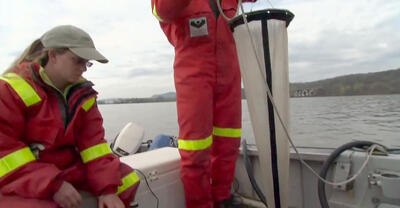 Two scientists, one standing and holding a long mesh net and one sitting, on a motorboat on the river.