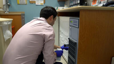 Dr. Ari Grinspan crouches in front of a freezer, holding a small box in his right gloved hand.