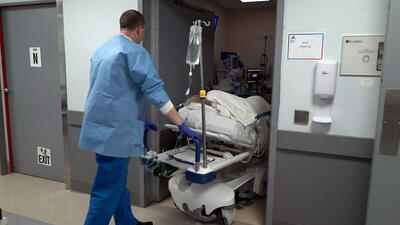 A doctor in a gown wheels a patient on a stretcher into a room.