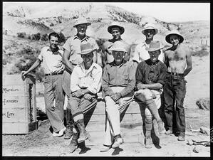 Barnum Brown sits flanked by two people and five additional people stand behind them, all wearing hats with mountains in the background.