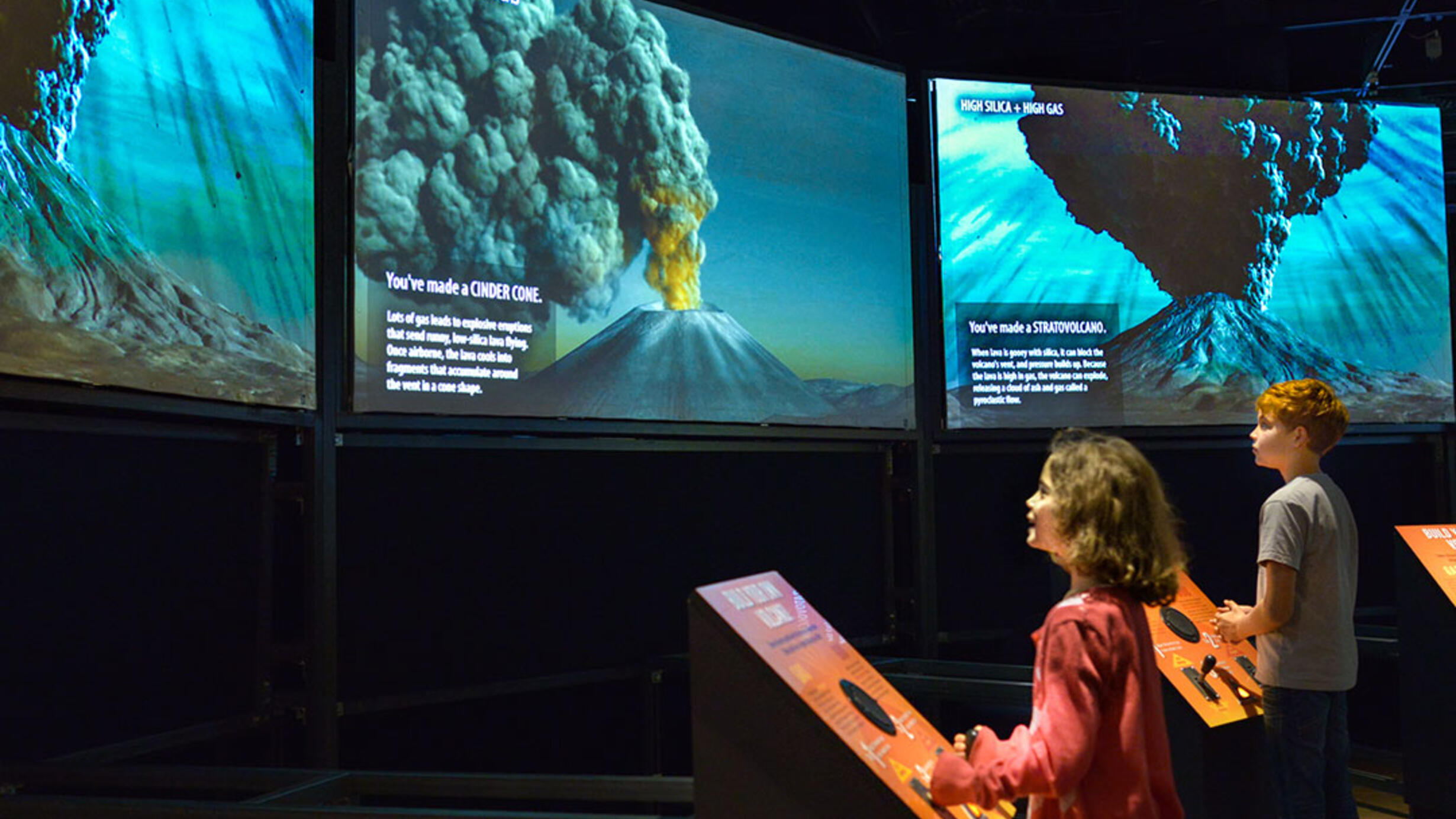 Two children in a museum hall looking at large wall screens showing different kinds of volcanoes erupting. They are interacting with “Build Your Own Volcano” touch screens.