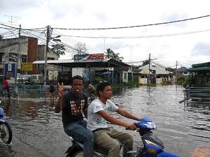 Two young men riding a scooter through a flooded street.