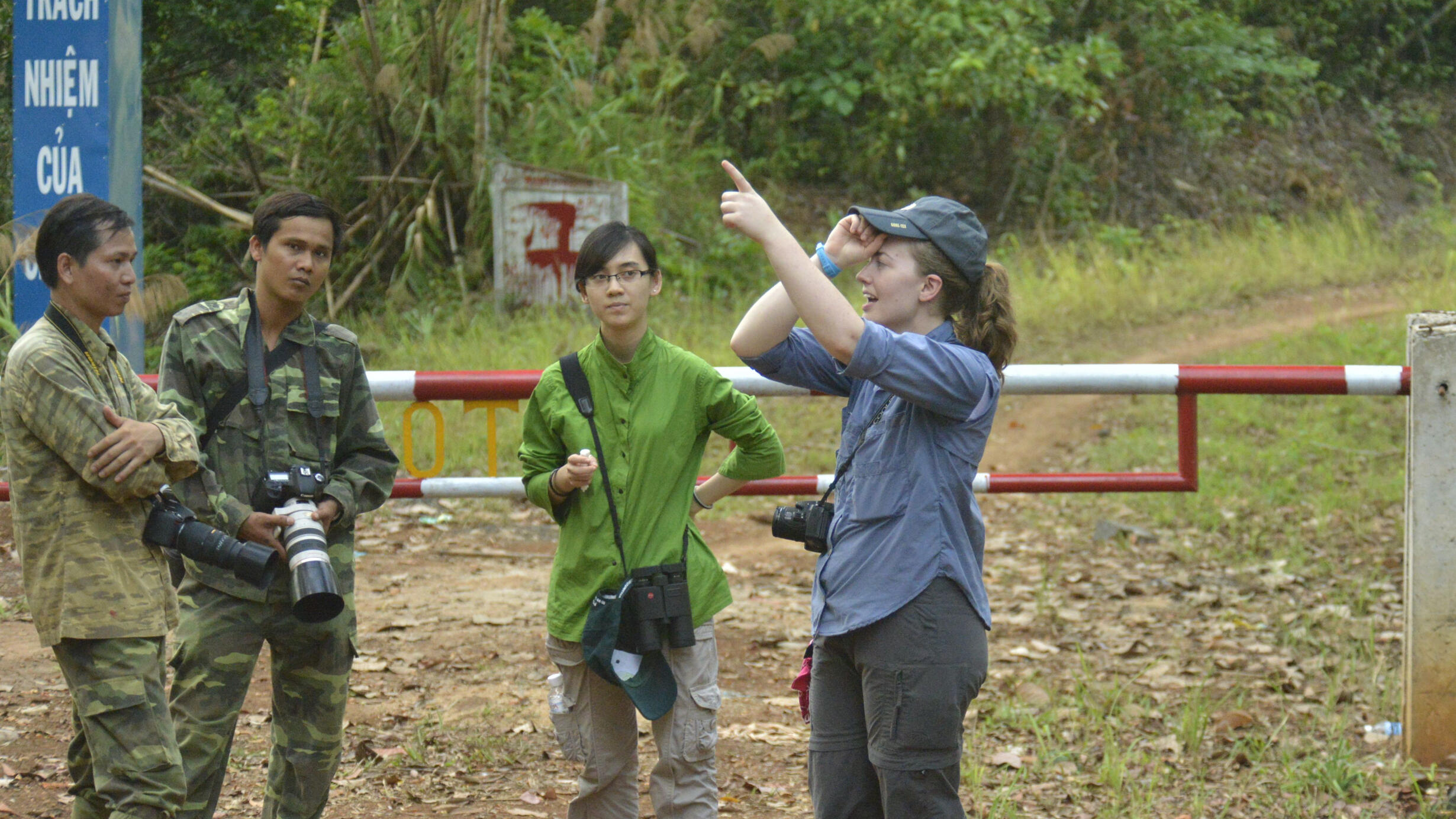 Four people with cameras around their necks, two in military fatigues and one pointing upwards, stand by a barrier arm and a sign in Vietnamese. 