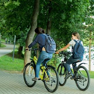 Two people wearing backpacks ride bikes along a paved path lined with grass and trees.