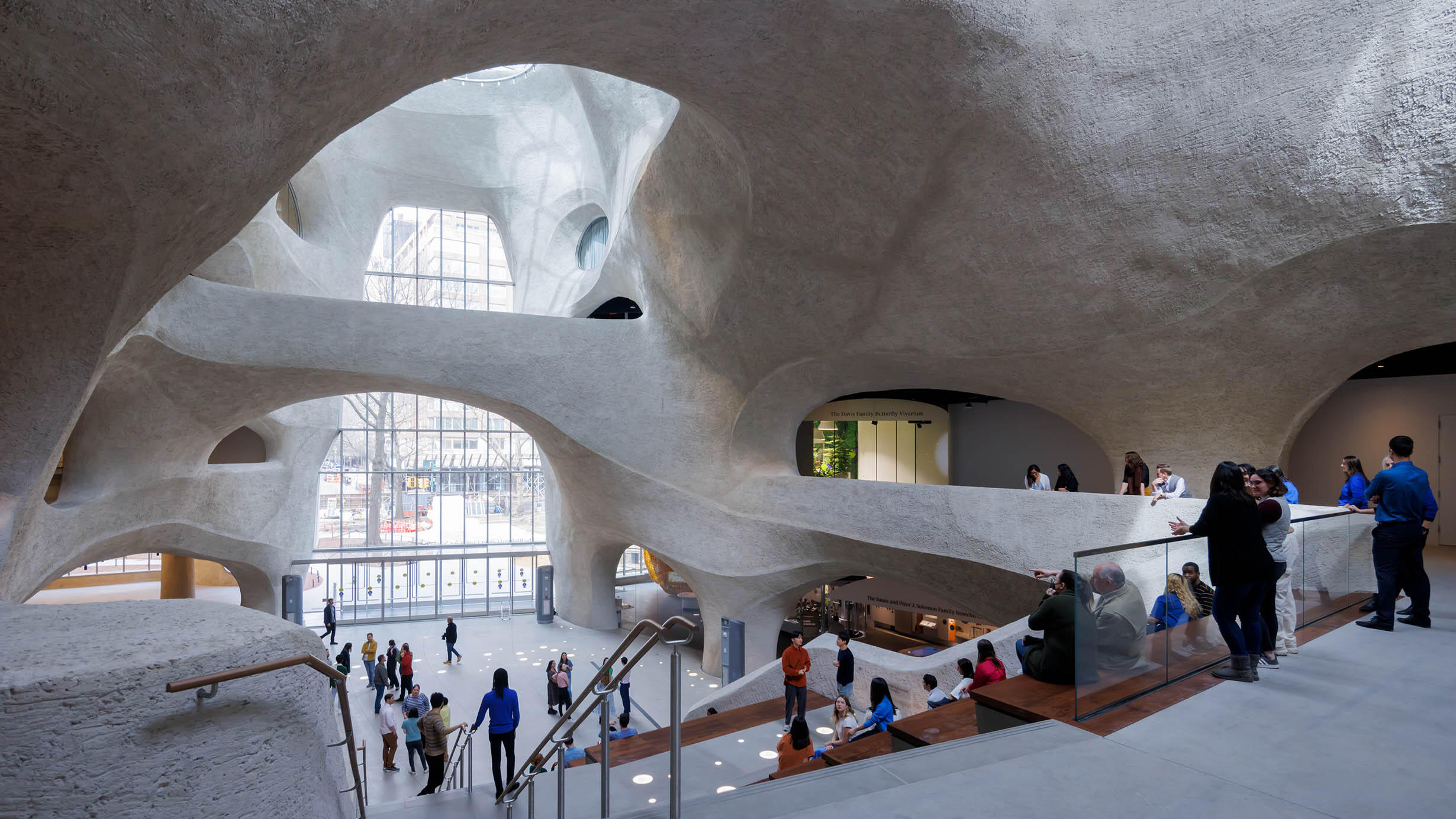 Museum visitors sit, stand, and walk around the second floor, and the stairs down to the exit, of the atrium inside the Richard Gilder Center.