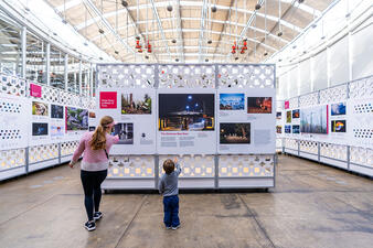An adult and child point at text and images on a free-standing wall in an exhibit installation in a large room.