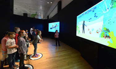 A group of children gesture in front of an interactive game that cause creatures to behave in ways consistent with some of their abilities