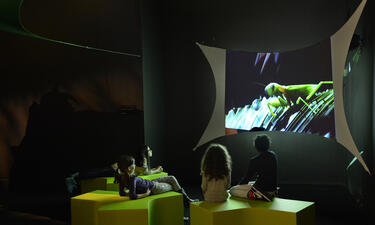 A group of children sit on green chairs in front of a giant screen displaying an image of a grasshopper