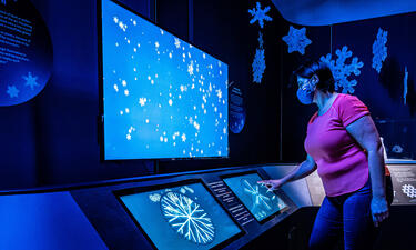 A woman in a red shirt points to a screen with falling snowflakes on it.