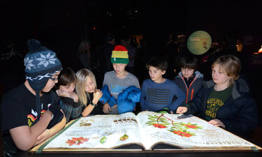 Siete niños alrededor de una gran libro iluminado con ilustraciones botánicas.
