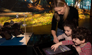 A woman and two small children surround a digital touchscreen that shows a T. rex illustration and tools for adjusting the animal's appearance.