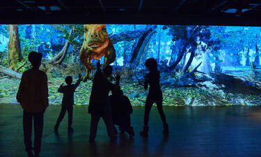 Silhouettes of a group of people of different ages are shown interacting with an animation of a T. rex in a forest that fills the entire wall