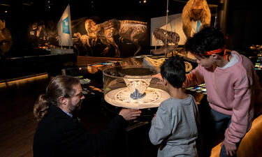 An adult and two children kneel down to look inside of a praxinoscope which shows movement through illustrations reflected in mirrors as it spins.