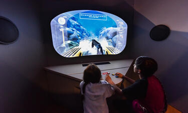 Two children use controllers as they sit in front of a monitor displaying an animated underwater scene