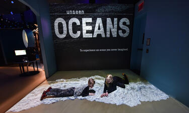 Two children lay on the floor on top of a projection of ocean waves on a beach, a wall behind displays the exhibition title Unseen Oceans