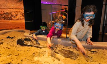 Three small children wear plastic protective goggles as they dig in a sand pit