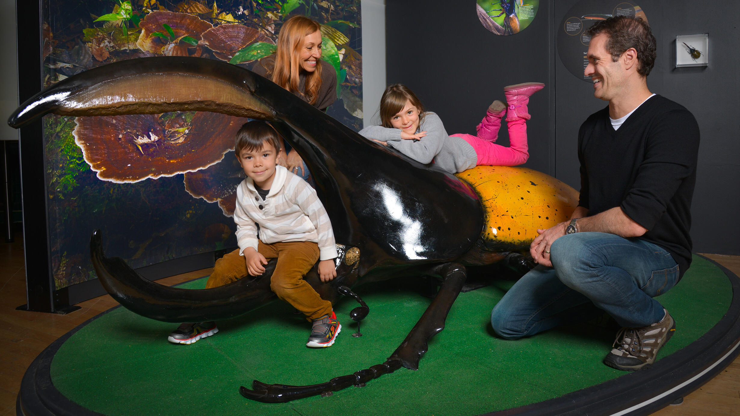 A young boy and girl climbing on a giant model of a hercules beetle while their parents watch