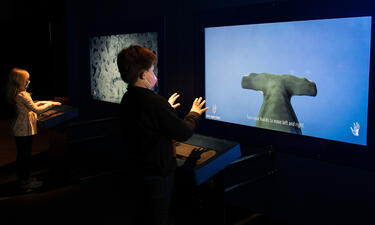 A boy stands in front of a screen with his hands out, piloting a hammerhead shark on the screen.