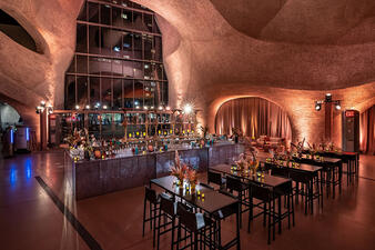 High tables and chairs are placed in front of a large horseshoe-shaped cocktail bar in the dramatically-lit Gilder Center atrium.
