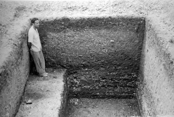 Black-and-white photo of a researcher standing inside an excavated rectangular hole.