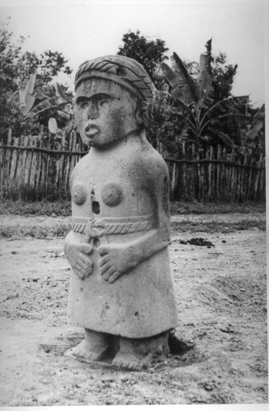 Carved stone column of a fertility goddess in a belted skirt. Bas-relief arms bend at elbows, hands resting near belly. Breasts are bas-relief circles.