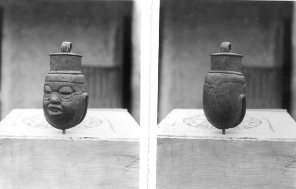 Black-and-white photos, front and back of an oval-shaped copper bell in the image of a human face. The base has an opening similar to a clamshell.