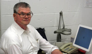 A man wearing eyeglasses smiling at the camera and seated at a desk with a computer and telephone.