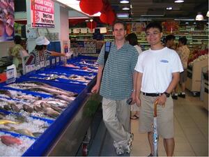 Two men posing for the camera in an indoor fish market with trays of fish displayed on ice.